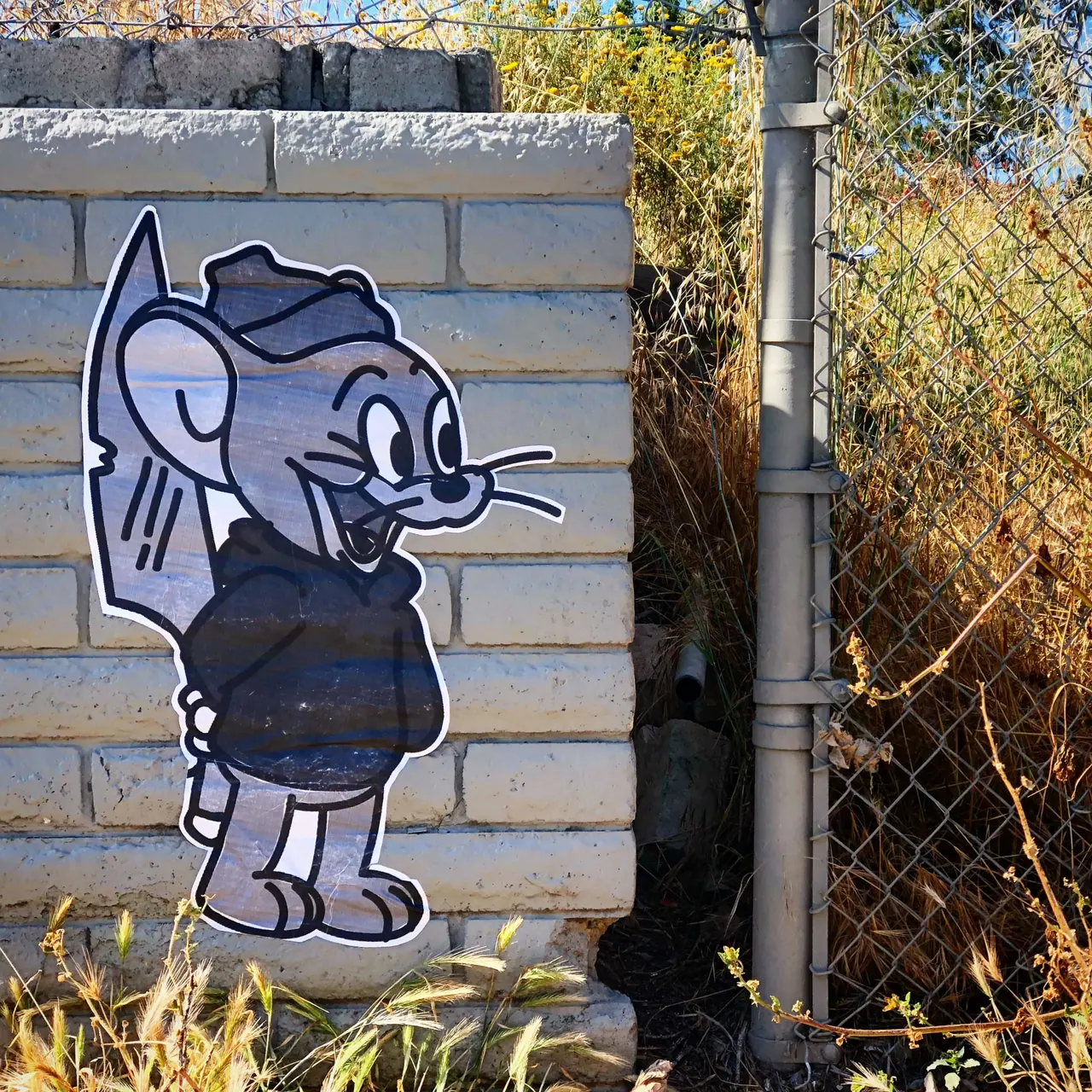 A cartoon mouse pasted onto a brick wall beside a chain-link fence.