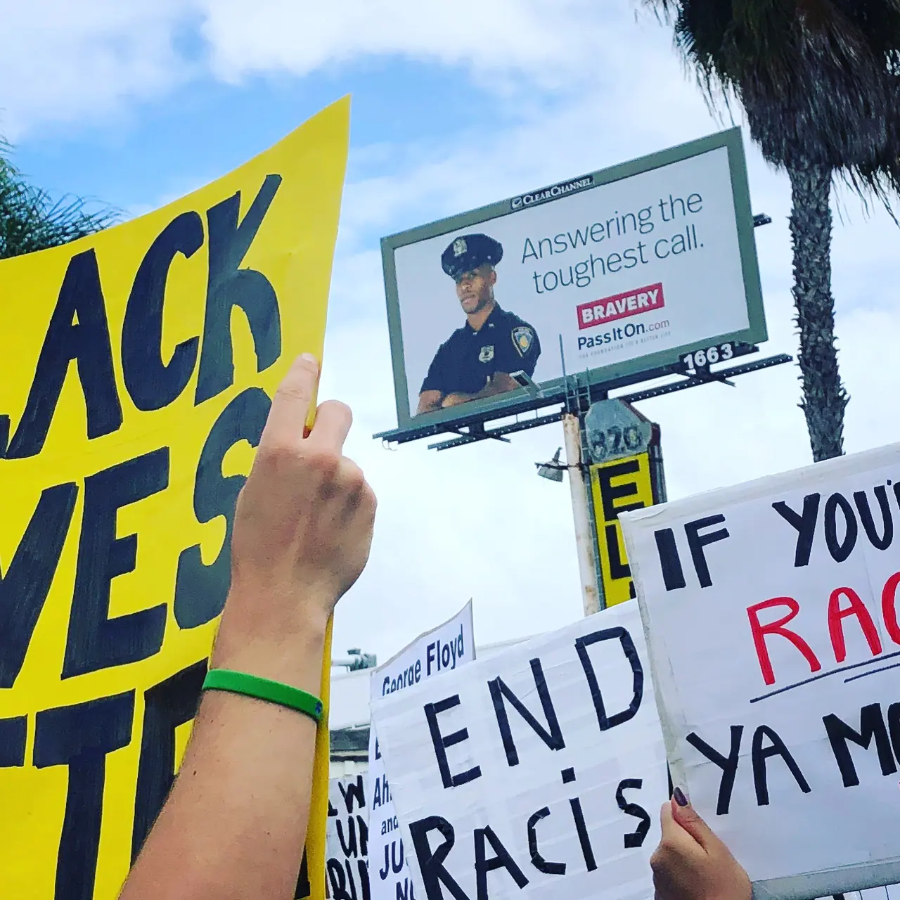 Raised protest signs beneath roadside billboards and palm trees.