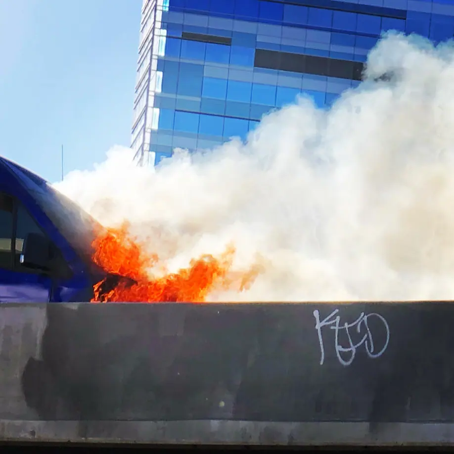 Flames and smoke rising from a truck beside a freeway barrier.