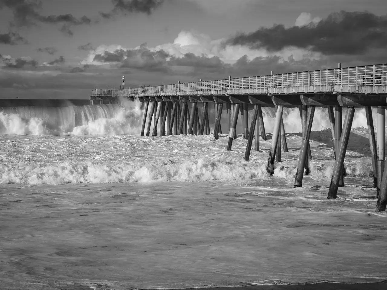 Large waves breaking against Hermosa Beach Pier, in black and white.