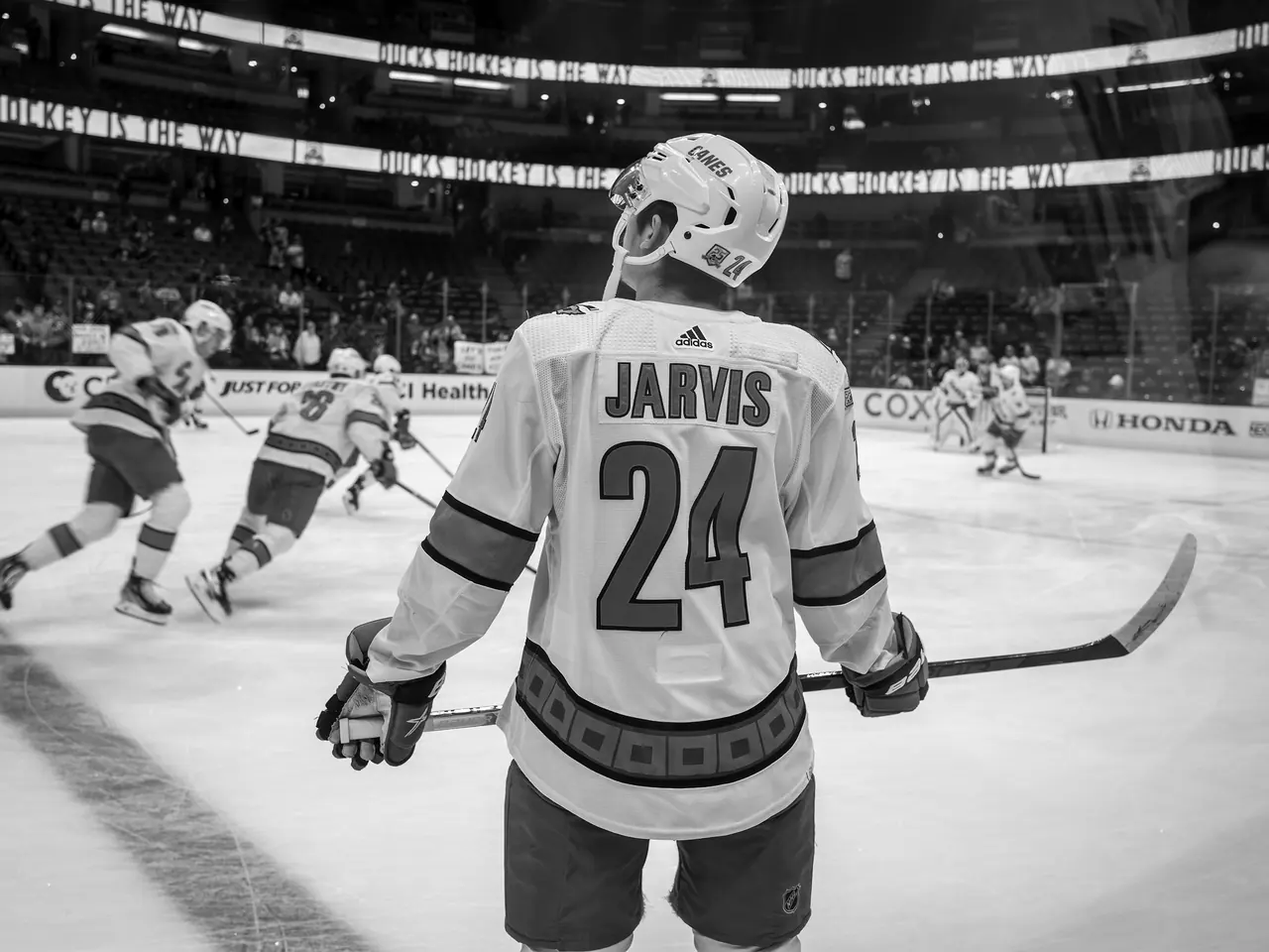 A hockey player watching teammates on the ice from beside the boards.